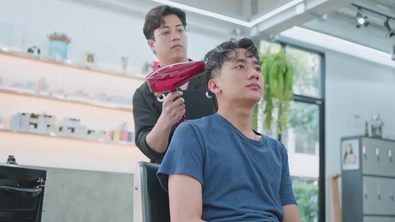 亚洲有魅力的专业男性发型师或发型师利用吹风机吹湿年轻男性顾客的头发，在美容院或理发店进行发型保养或职业服务。