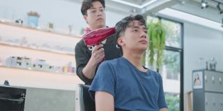 亚洲有魅力的专业男性发型师或发型师利用吹风机吹湿年轻男性顾客的头发，在美容院或理发店进行发型保养或职业服务。