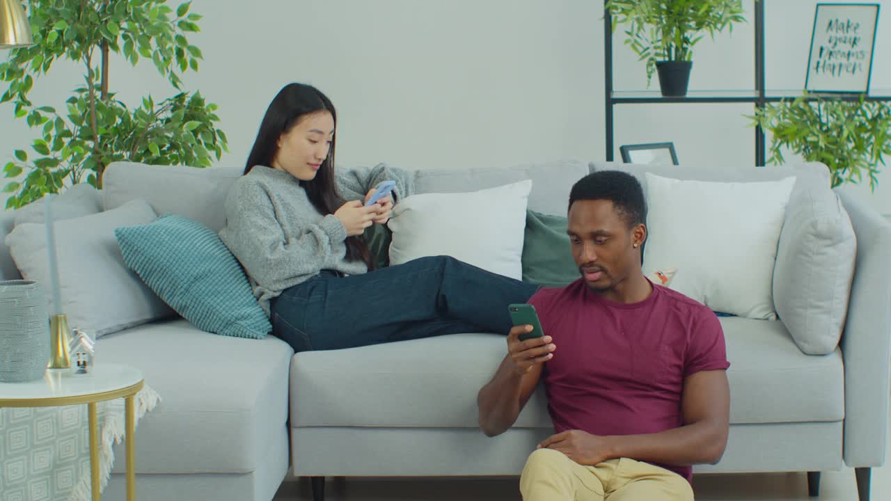 年轻的跨国情侣，无视对方看手机。男人和女人躺在沙发上用着智能手机。