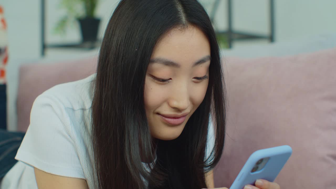 漂亮的亚洲女人躺在客厅的沙发上拿着智能手机，聊天使用社交媒体网络，观看来自朋友的视频。