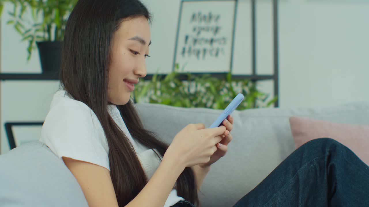 快乐专注的混血儿年轻女子聊天用智能手机坐在舒适的沙发上。亚洲现代女性上网或在电话上打字。