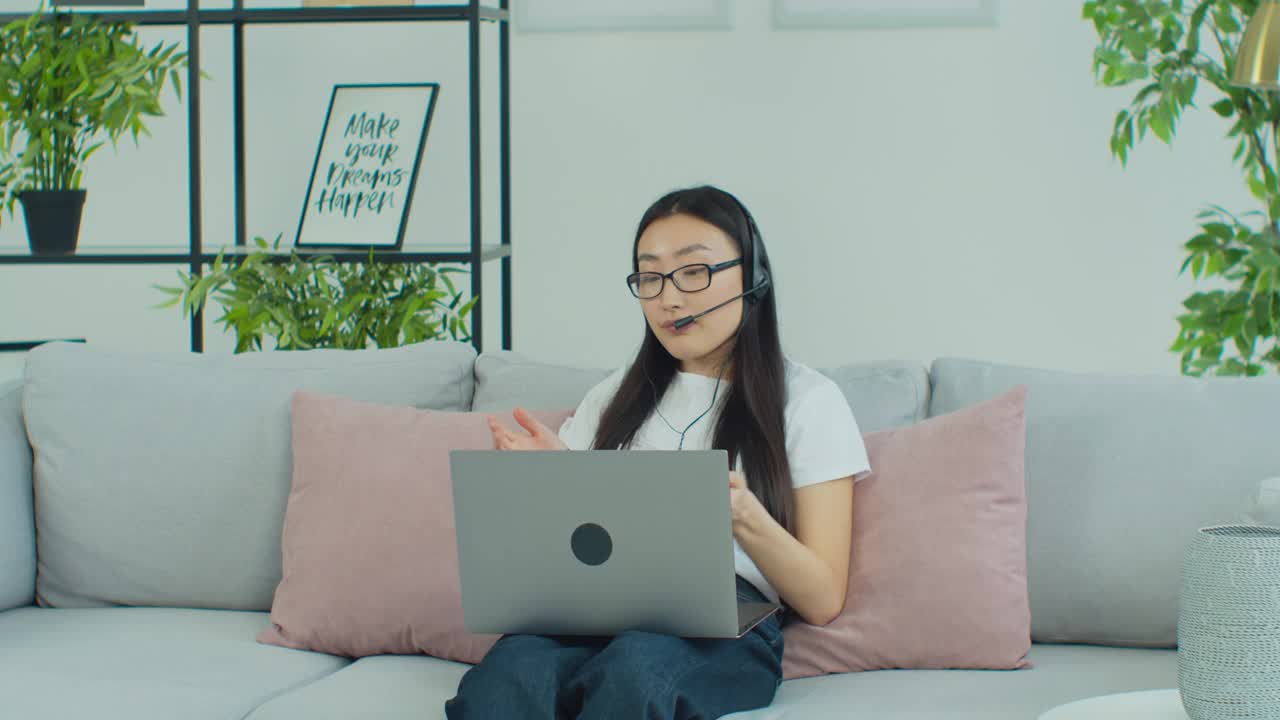 年轻的亚洲女性戴着耳机在笔记本电脑上开会，与在线教师聊天，学习，在家工作。亚洲混血女学生在眼镜e学习用电脑摄像头聊天做笔记。远程教育的概念。
