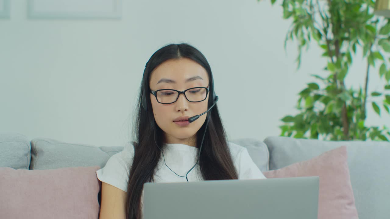 年轻的女孩学生戴着眼镜戴着耳机学习通过网络视频聊天在家。千禧女性在笔记本电脑上召开视频会议，通过网络摄像头做记录。远程教育的概念。