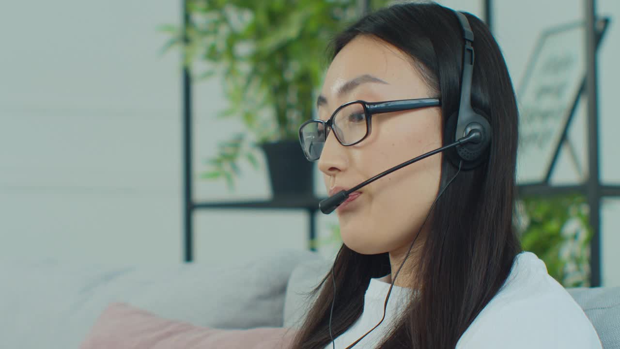 亚洲妇女戴眼镜呼叫中心话务员，客户支持员戴耳机在笔记本电脑上咨询客户在线打字，在网上聊天，服务电话接线员秘书进行视频会议。