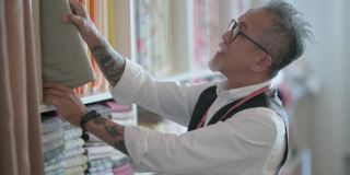 面部留胡茬的亚洲华人成熟男子裁缝在工作室检查他的织物服装