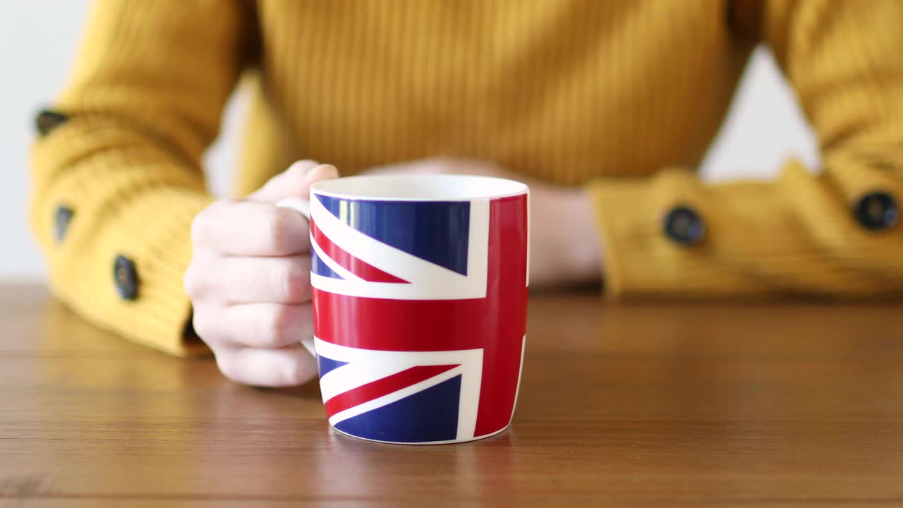 特写的手在一个木制的桌子上一个英国国旗杯热气腾腾的饮料，被一个年轻的女人举起