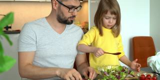 父亲和小女儿在厨房的餐桌上准备蔬菜、莴苣叶和西红柿沙拉。小女孩帮忙做饭。