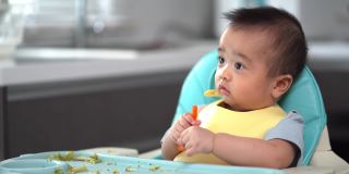 宝宝看了看手里的胡萝卜。第一次吃泥。他的亚洲小男孩的母亲喂养婴儿食品。