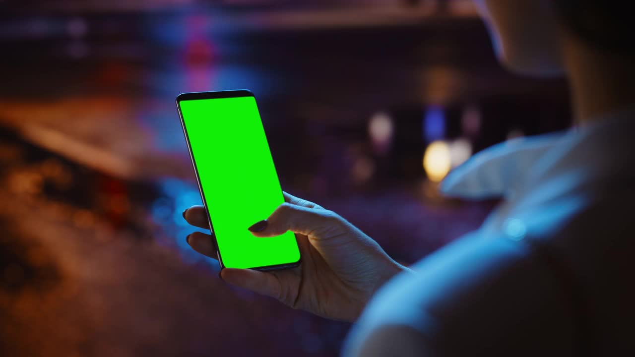 女人站着使用Chroma Key智能手机，打手势，滑动，滚动。霓虹夜市大街。女性使用绿屏手机。肩部特写镜头