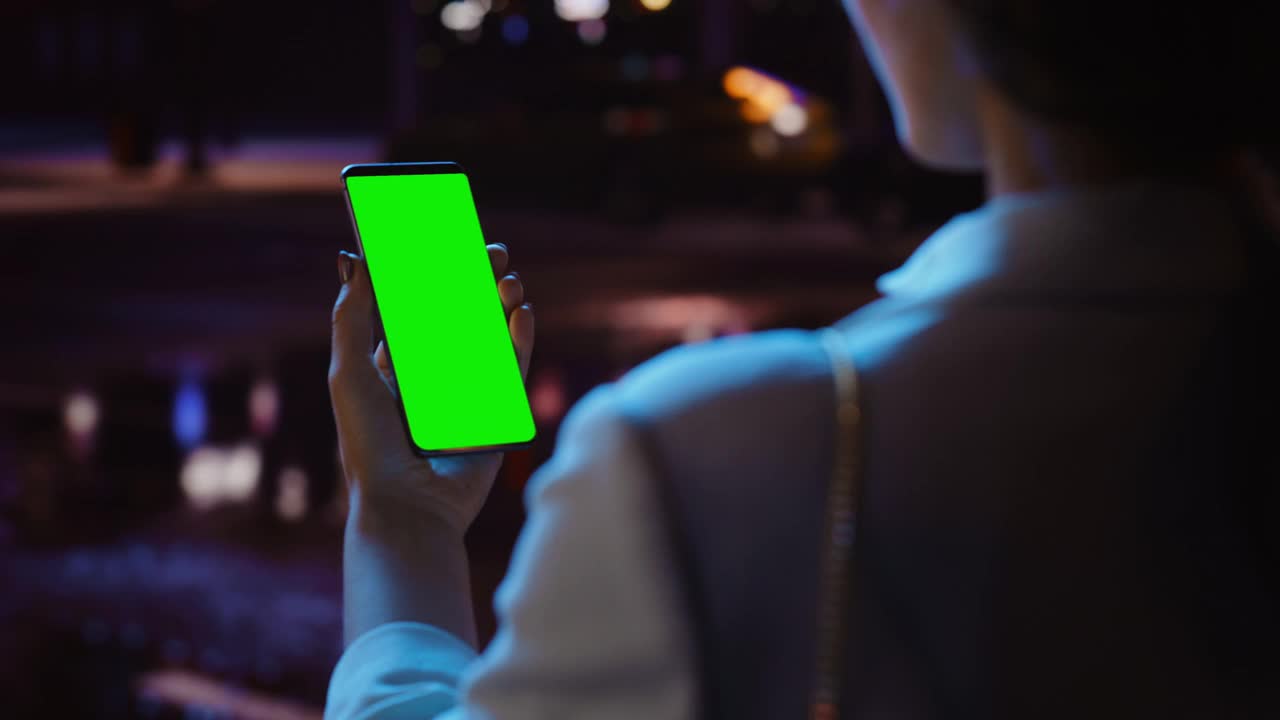 站着看绿屏的女人用Chroma Key智能手机霓虹夜市大街。使用手机的女性。过肩特写跟踪镜头聚焦显示