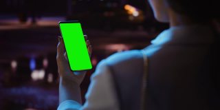 站着看绿屏的女人用Chroma Key智能手机霓虹夜市大街。使用手机的女性。过肩特写跟踪镜头聚焦显示