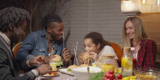 快乐的多民族的人们坐在户外的餐桌上吃着野餐汉堡，有说有笑。愉快的白人妇女非裔美国男人和女孩在院子里聊天在黄昏。缓慢的运动。
