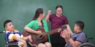 亚洲美女弹吉他鼓励青少年唐氏综合症女孩练习弹吉他，跳舞，唱歌与残疾学生团体在轮椅上音乐会议课感受快乐，积极的情绪。
