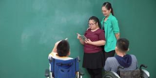 十几岁的唐氏综合症女孩练习演讲，在一个数字平板电脑上读演讲，残疾学生群体坐在轮椅上，亚洲美丽的女教师站在课堂上给予鼓励，感到幸福，积极的情绪。