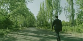 后视镜:西装革履的人独自穿过森林或花园的树木。孤独的男孩走在林间的小径或人行道上，林间有高大的绿树。缓慢的运动。无人机
