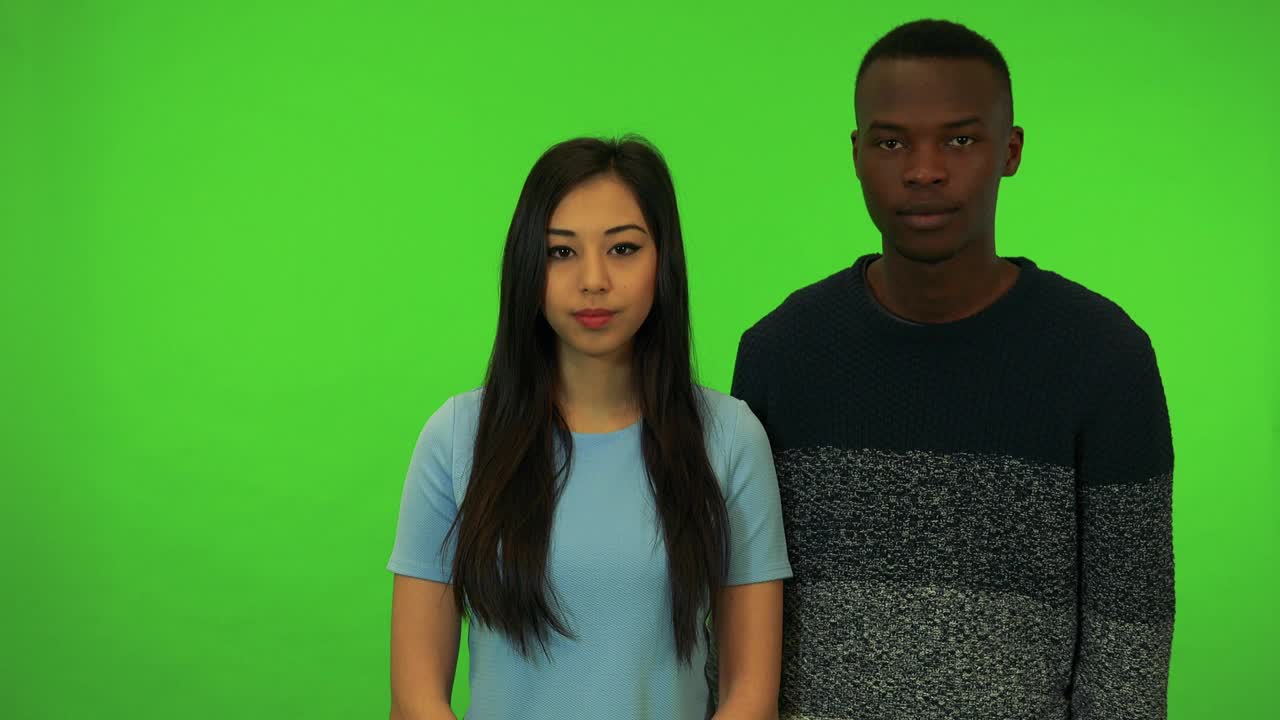 一个年轻的亚洲女子和一个年轻的黑人男子微笑着向镜头挥手——绿幕摄影棚
