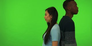 一个年轻的亚裔女子和一个年轻的黑人男子背对着对方站在一起，面带微笑地对着镜头——绿幕摄影棚