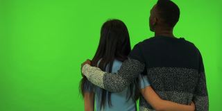 一个亚洲女人和一个黑人男人用他们的手臂环抱着对方和他们的背对着镜头看东西-绿色的屏幕工作室