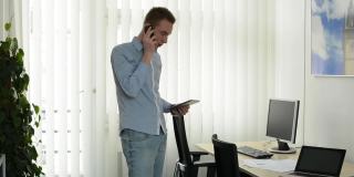 年轻英俊的男子用智能手机打电话，在办公室用平板电脑工作，看着窗外