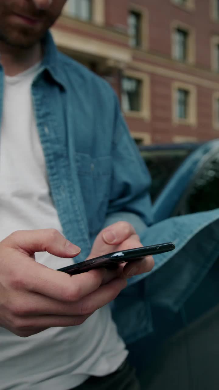 一个穿着牛仔裤衬衫的男人靠在一辆蓝色汽车上打手机的特写镜头