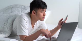 一名年轻的亚洲男子躺在床上使用智能手机和笔记本电脑