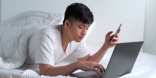 一名年轻的亚洲男子躺在床上使用智能手机和笔记本电脑