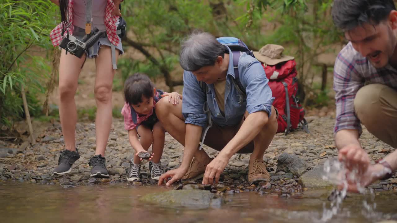 一个亚洲大家庭带着一个孩子，坐在小溪边休息，爸爸，爷爷，妈妈洗着汗，女儿和爷爷奶奶玩得很开心，心情激动，在热带森林探险和徒步旅行。