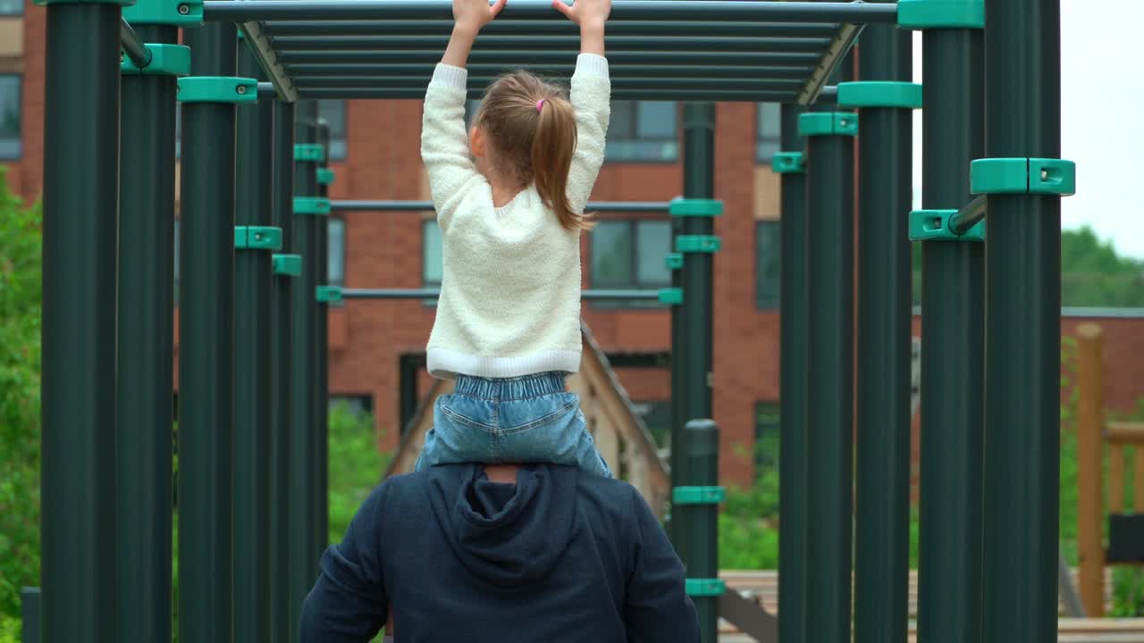 爸爸和小女孩在户外操场上运动的背影。快乐的家庭，小女孩和爸爸在绿色的夏天公园里玩活跃的游戏。