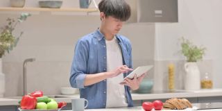 年轻的亚洲男子站在厨房里使用平板电脑