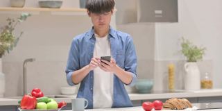 年轻的亚洲男子站在厨房使用智能手机