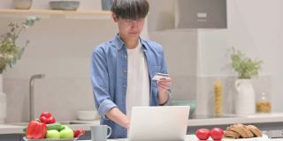 年轻的亚洲男子在厨房用笔记本电脑在线支付