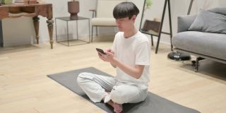 年轻的亚洲男子坐在瑜伽垫上用智能手机聊天