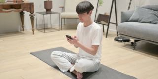 年轻的亚洲男子在瑜伽垫上使用智能手机