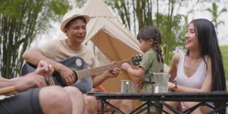 一旁看亚洲的爷爷、爸爸、妈妈、女儿心情愉快地弹着吉他，一起唱着歌。冒险多代家庭露营，社交聚会与帐篷，周末户外活动。