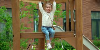小女孩坐在操场上的滑梯上，看着相机，在绿色的夏日公园里笑个不停。快乐的孩子在户外玩耍。
