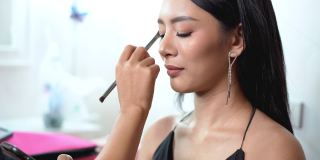 肖像美丽的亚洲女人自然化妆与许多亮和她使用手机。化妆师用刷子把眼影刷到眼睛上。闪亮的眼影