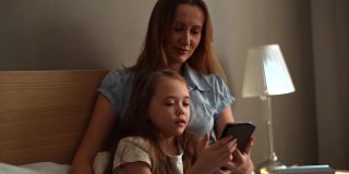 快乐的年轻母亲拥抱学龄前的女儿一起浏览和上网手机的特写。