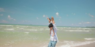 夏天的海滩上，亚洲婴儿坐在他爸爸的脖子上。父母和孩子的角色扮演和幻想是飞机和飞行员。幸福家庭和旅游理念。