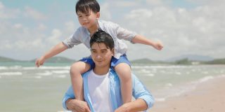 一个亚洲人的儿子夏天在海滩上坐在他父亲的脖子上。父母和孩子的角色扮演和幻想是飞机和飞行员。幸福家庭和旅游理念。