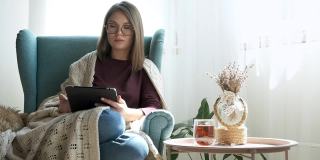 年轻女子戴着眼镜使用数码平板电脑，喝着茶，坐在家里的扶手椅上