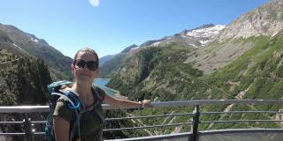 一名戴着墨镜的妇女站在奥地利卡林西亚的科隆布雷恩大坝旁。大坝被高山环绕着。陡峭的斜坡。那个女人微笑着，享受着阳光明媚的日子。