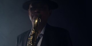 优雅的男子在一顶帽子和正式的西装演奏萨克斯在深灰色的背景在烟雾演播室。音乐家在黑暗中吹萨克斯管