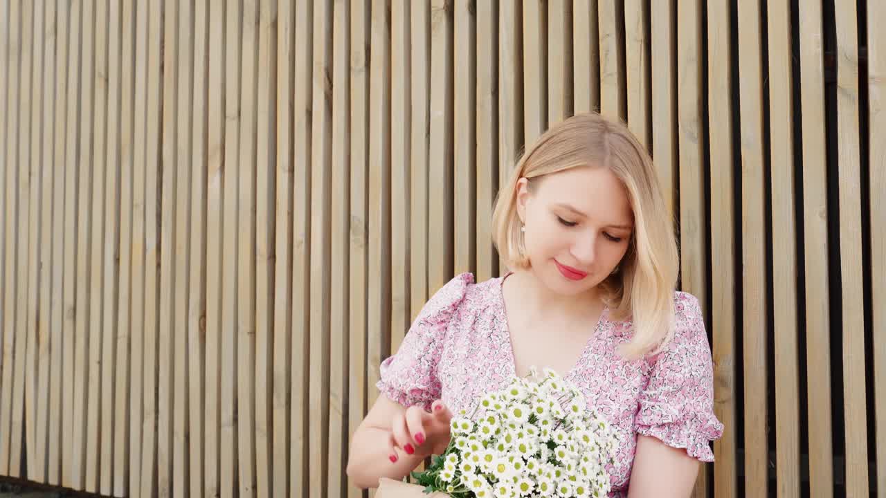 金发短发，涂着红色唇膏的时髦女人在户外放松。可爱的女孩与一束洋甘菊和纸袋法国法棍。看着镜头，开玩笑地扯下花瓣，吹着风