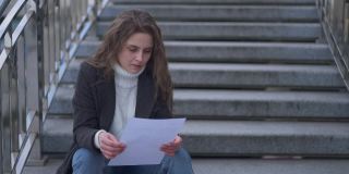 年轻的黑发白人悲伤的女人读信撕纸坐在城市的楼梯户外。压抑压抑愤怒的千禧年春日肖像秋日。缓慢的运动。