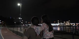 跟踪跟踪摄像头拍摄的两个快乐的姐妹笑着微笑着走在海滨地区的步行街在灯泡下在夏天的夜晚，一位女士把她的手臂放在另一个女人的肩膀4K分辨率