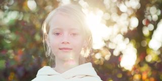 美丽的金发小女孩在白色帽衫看自信的相机摆姿势在户外的阳光，树木的叶子散景在背景