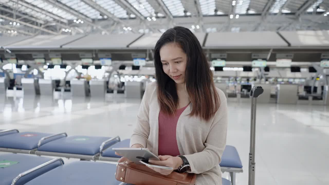 一名亚洲女子身穿粉色t恤，带着行李、护照、机票等在值机柜台前的椅子上，用数码平板电脑检查航班时间，挥手微笑，在机场开始一段新的旅程，旅行，度假。
