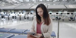 一名亚洲女子身穿粉色t恤，带着行李、护照、机票等在值机柜台前的椅子上，用数码平板电脑检查航班时间，挥手微笑，在机场开始一段新的旅程，旅行，度假。