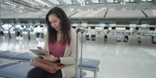 一名亚洲女子身穿粉色t恤，带着行李、护照、机票等在值机柜台前的椅子上等朋友，她在机场用电子平板电脑查询航班时间，开始一段新的旅程，旅行，度假。