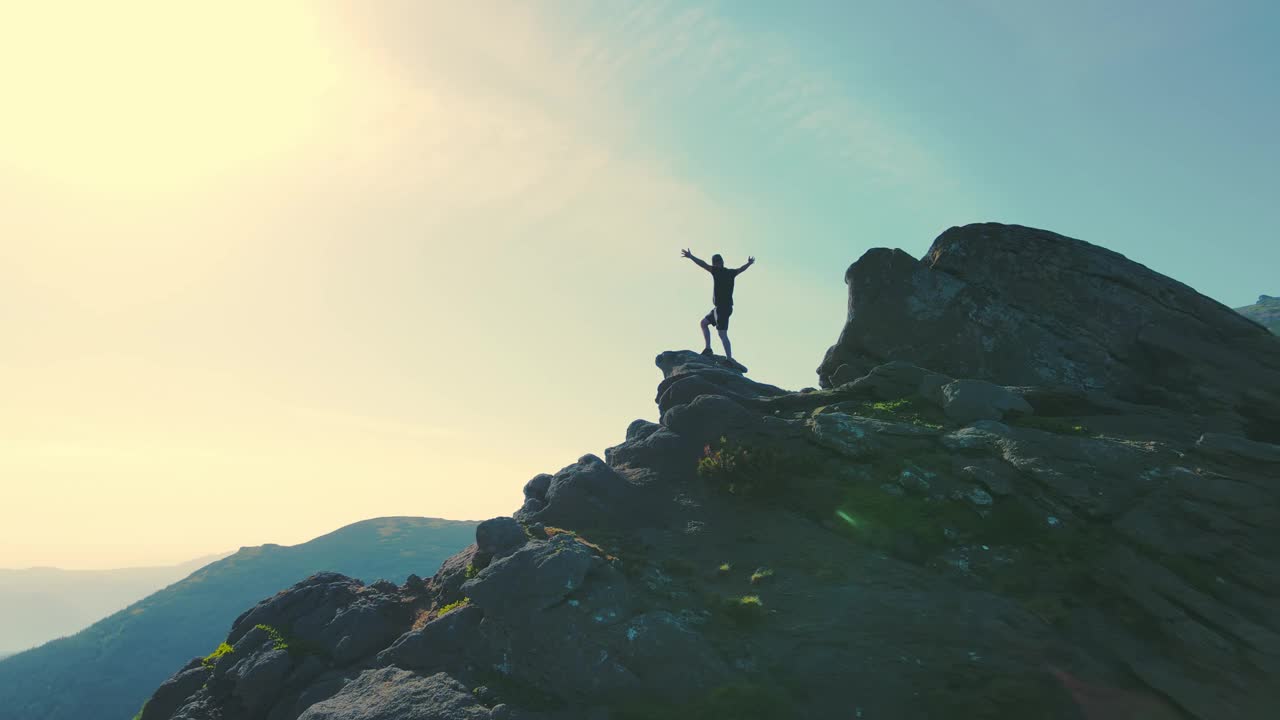 Aeria绕着一个站在山顶的男人飞着，他举起双手望向远方。一个游客的剪影在岩石上看未来在日落